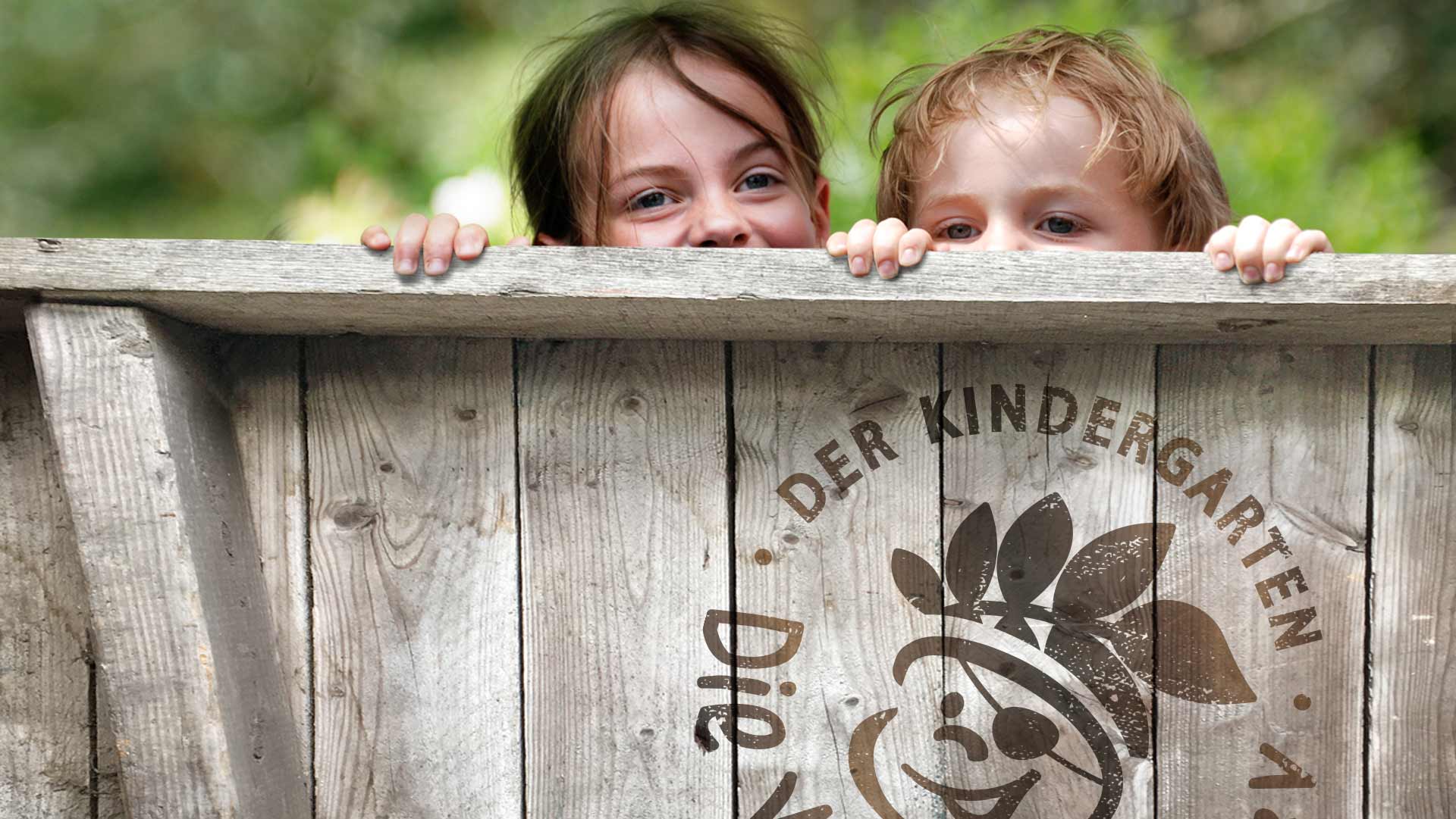 Zwei Kinder, die über eine Holzwand lugen, auf der Holzwand ein großes eingebranntes Logo